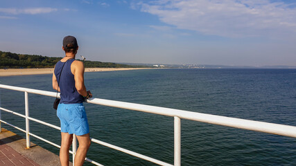 A man in shorts and full-cap standing at the pier by the Baltic Sea side in Poland. There is a sandy beach stretching along the costal line and a dense forest behind it. Calm sea. Serenity