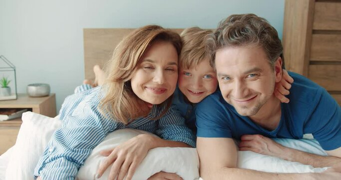 Portrait of happy Caucasian family: mom, dad and little son lie in bed in the morning resting and smiling looking at camera.
