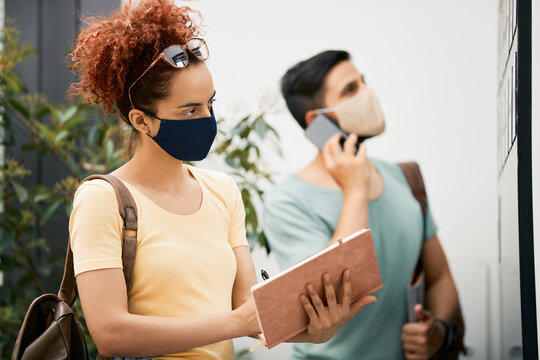 Female Student With Face Mask While Reading Announcements On Noticeboard At University Hallway.