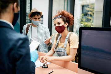 Smiling female tourist wearing face mask while communicating with receptionist and checking in a...