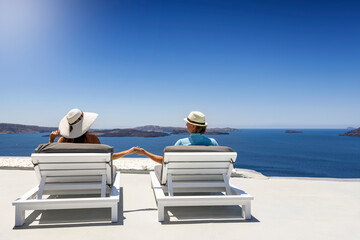 A romantic tourist couple sits holding hands on white sunbeds and enjoys the summer view over the...