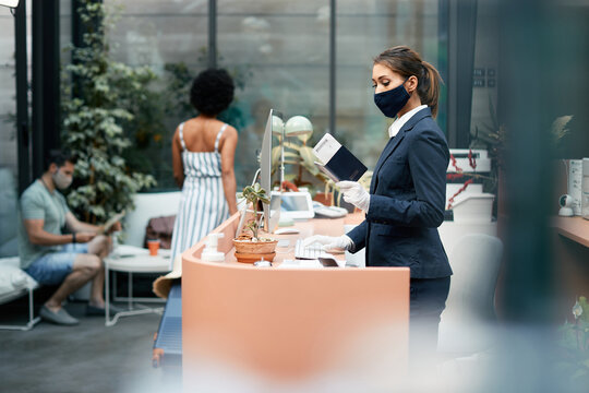 Hotel Receptionist With Protective Face Mask Entering Guest's Data On A Computer While Reading Their Passport At Reception Deck.