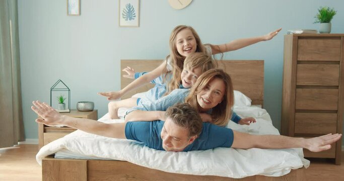 Portrait of happy Caucasian family lying together on bed on top of each other balancing and spreading their arms to side depicting airplane.
