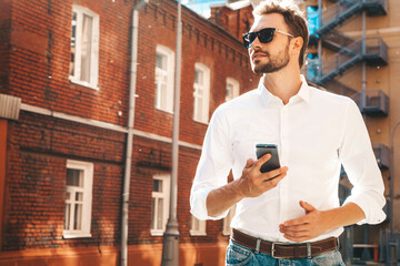 Fototapeta na wymiar Handsome smiling stylish hipster lambersexual model.Modern man dressed in white shirt. Fashion male posing on the street background in sunglasses. Outdoors at sunset. Using smartphone apps
