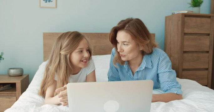 Happy Caucasian mother and daughter lying together at home on bed and watching cartoons on laptop having fun.