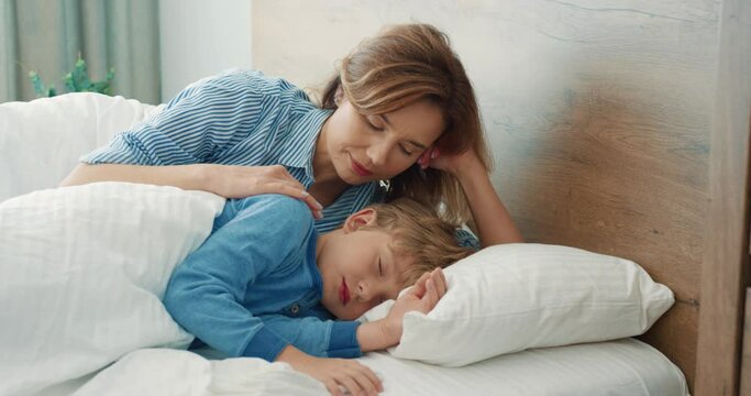 Loving mother lies in bed in morning with her little sleeping son and looks at him stroking. Maternal tenderness.