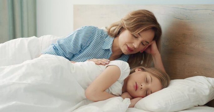 Happy Caucasian mother lies next to her sleeping daughter in bed and tries to gently wake her in the morning.