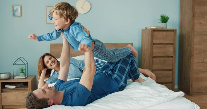 Happy Mom And Dad Cheer Up Little Son Lying In Bed In Morning And Holding Him In Air Like Airplane.