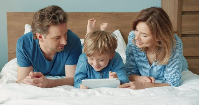Happy father and mother spend their free time with their little son lying in bed watching cartoons on tablet.