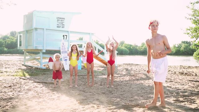 Cute blond teen boy with red headband and white swim shorts jumping at the sand beach against blue lifeguard tower with his friends.