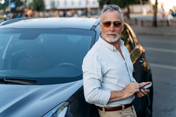 Senior man standing near his SUV car and holding smartphone. Middle aged man portrait outdoors near...