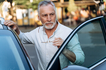 Senior man is standing near his rental SUV car and holding sunglasses. Middle aged man portrait...