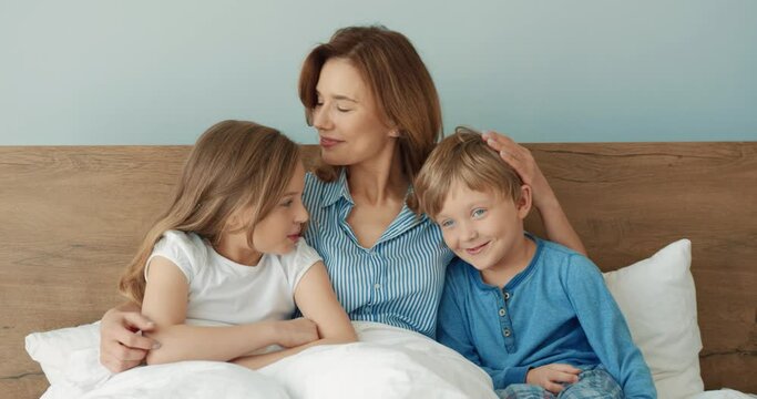 Happy beautiful mother sitting in bed with daughter and little son hugging and giving them her tenderness and care.