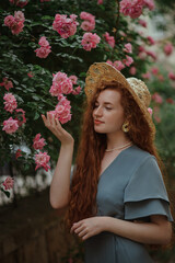 Beautiful redhead freckled woman with long curly natural hair, wearing straw hat, blue dress, posing in blooming rose garden, touching flowers. Beauty, hair care, summer lifestyle concept. 