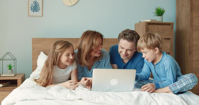 Happy parents with two children spend free time together lying at home on bed watching comedy on laptop.