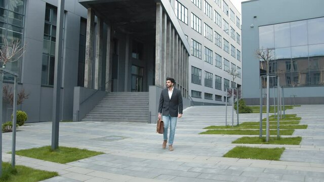Attractive businessman walking outside near big office building and looking around. Muslim businessman in stylish formal wear with brown leather suitcase in hands. Bearded young guy on way to his work