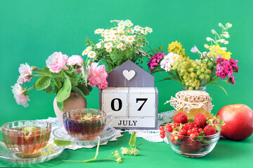 Calendar for July 7 : the name of the month of July in English, cubes with the numbers 0 and 7, bouquets of wild flowers, jam, fruit, cups of tea, white napkins, green background