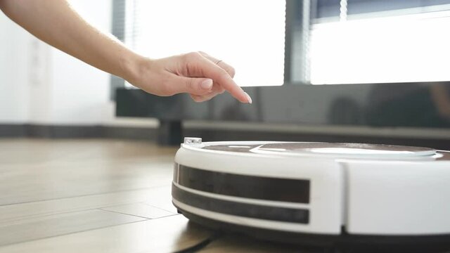 Female push start button on robot vacuum cleaner. Modern smart household. Detail of hand that turns on smart vacuum robot pushing its button. Robotic vacuum cleaner on floor in living room.