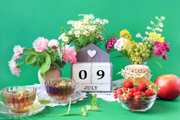 Calendar for July 9 : the name of the month of July in English, cubes with the numbers 0 and 9, bouquets of wild flowers, jam, fruit, cups of tea, white napkins, green background