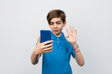 happy pretty boy with smartphone having video call and waving hand, standing against light gray background