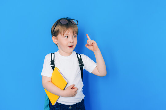 Back to school concept. Schoolboy boy with backpack and holding yellow book in front of blue background. Child shows thumbs up