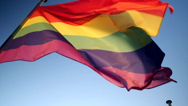 Slow motion shot from below a pride flag waving in the wind, clear blue sky background.