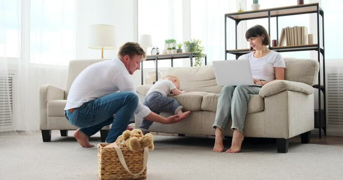 Baby son trying to climb sofa with help of father while mother is working on laptop at home