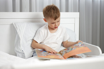 The boy looks with interest at the colorful pictures in his new book sitting in a white bed