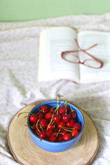 Bowl of fresh cherries, open book and glasses on a bed. Selective focus.