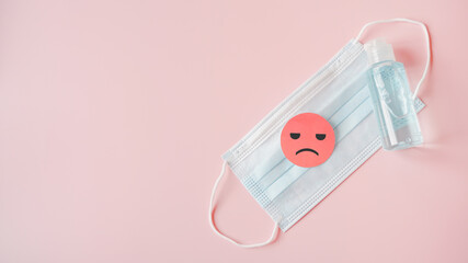 unhappy face on pink paper cut on medical face mask and bottle of blue alcohol gel on sweet pink...
