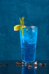 Fresh BLUE LAGOON summer cocktail on curacao background, with melted ice around it, garnished with lime, rosemary and cranberries.