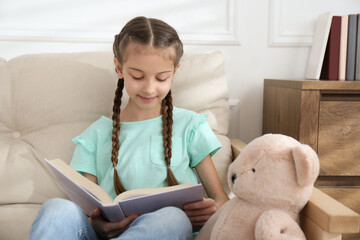 Cute little girl reading book on sofa at home