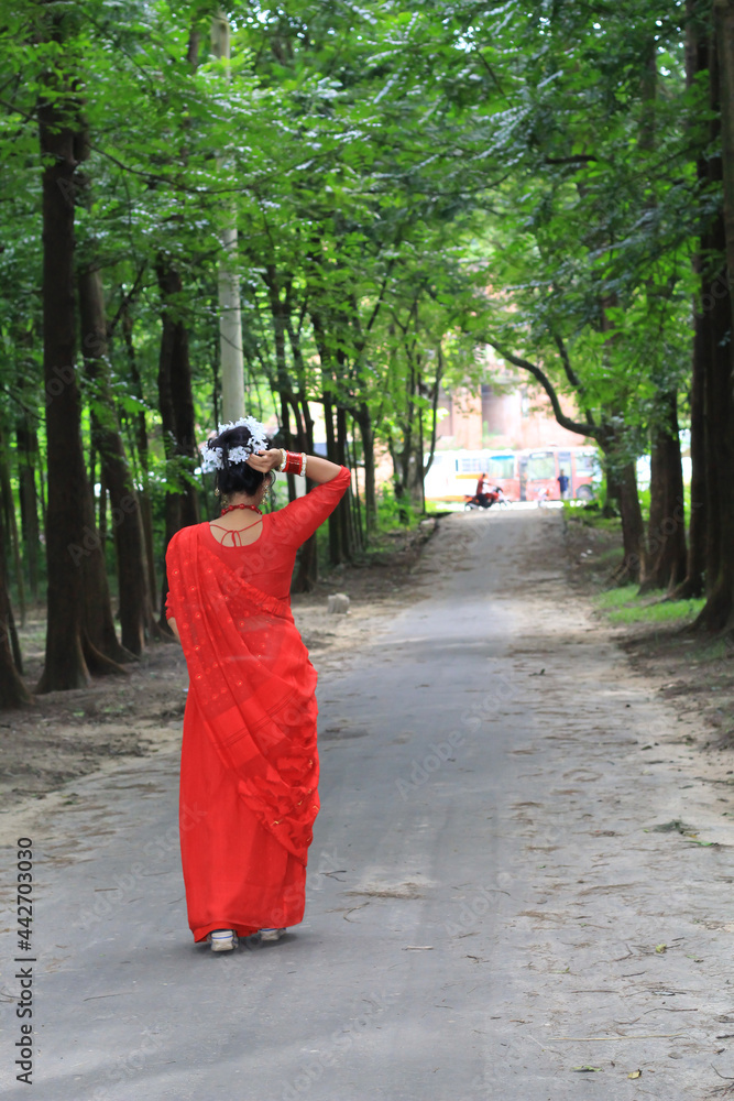 Wall mural young woman wearing a red saree walking on green asphalt road. the girl walks along the path in the 