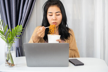 busy Asian woman eating noodles while working on laptop at office desk 