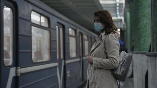 Woman wearing mask with backpack on shoulder standing on subway platform, train arriving. Commuters entering carriage. Public transport during pandemic. Concept of safety