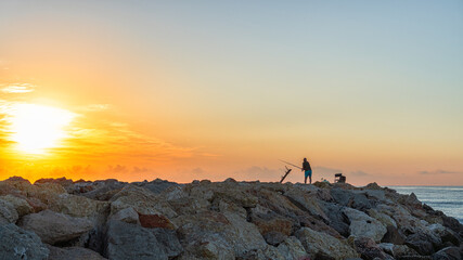 Man fishing on the breakwater at sunrise, near a port.