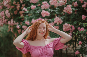 Happy smiling redhead freckled woman with long natural curly hair. Model posing in blooming roses garden, wearing pink headband, blouse. Summer lifestyle, fashion, beauty concept. Copy space for text