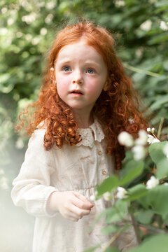 A beautiful little girl with red curly hair in a light dress, walking outdoors in a city park, looking at the sky. Portrait of a small child.