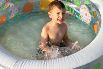 Little boy splashes in an inflatable pool. Squints from the spray. Backyard vacations concept