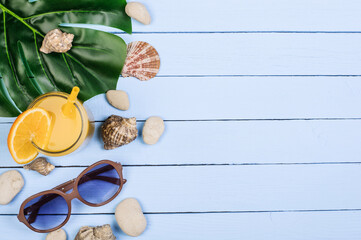 Glass with juice, sunglasses, seashells, monstera leaf on blue boards