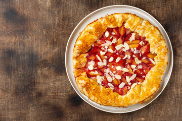 French Galette or a pie with red plums and almonds in a ceramic plate on the bright kitchen table top view. Summer traditional homemade sweet pastries