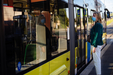 Lifestyle photo of middle age mature woman passenger in protective mask waiting while opening door to tram. Portrait of female alone go to work with bag on tram stop outdoor at summer morning.