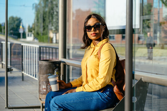 Casual black african woman in glasses sitting at the bus stop. Passenger waiting for city bus.