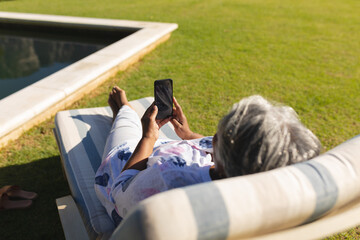 Senior african american woman using smartphone in deckchair by swimming pool in sunny garden