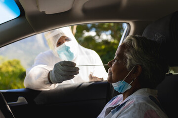 Medical worker wearing ppe suit taking swab test of senior african american woman sitting in car