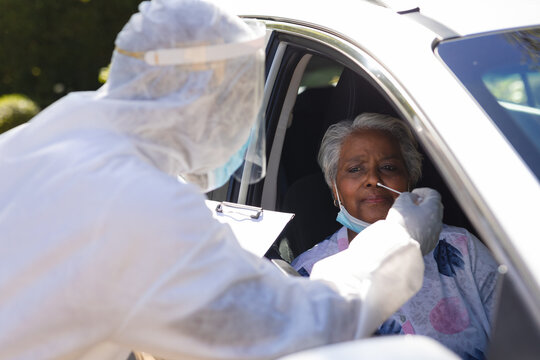 Medical worker wearing ppe suit taking swab test of senior african american woman sitting in car
