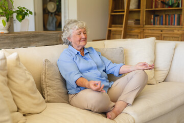 Senior caucasian woman sitting on sofa meditating with eyes closed