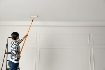 Young woman painting ceiling with white dye indoors, back view. Space for text