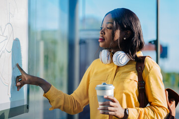 African female student in yellow blouse looking at of map public transport routes while waiting bus at stop at city street.