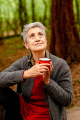 Mature lady with gray hair holding a cup of coffee with thoughtful gesture looking up in a forest. Feeling good in old age.